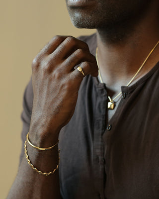 A close-up lifestyle shot of a man wearing gold jewelry, including a gold ring, a gold chain necklace with a small pendant, and a gold bangle bracelet, against a warm beige background.
