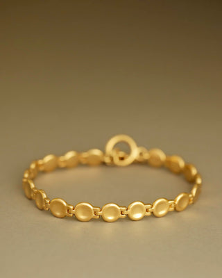 A gold link bracelet with flat circular disc links and a matching oval clasp, displayed on a neutral warm gray background.