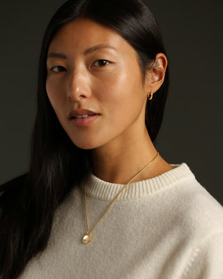 A portrait of a Chinese woman in a cream wool crewneck sweater wearing a polished gold pendant necklace with a smooth, oval disc pendant hanging from a gold chain.