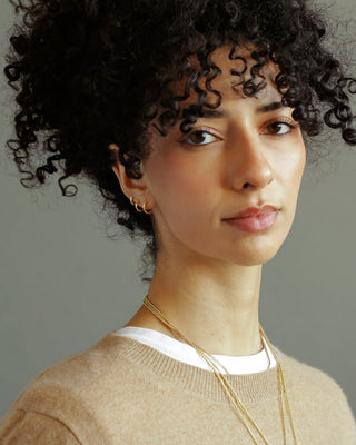 Woman with curly hair wearing a beige sweater and gold necklace against a gray background