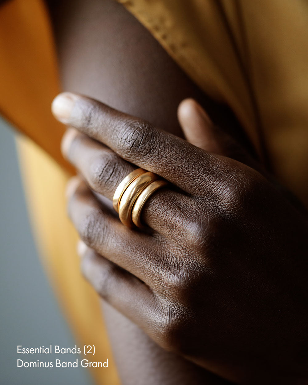 Black woman in silk dress wearing a stack of three solid 18k yellow gold simple women's wedding bands. Essential band and Dominus Band Grand by George Rings.
