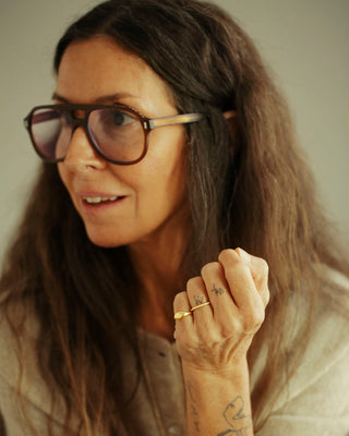A close-up portrait of a woman with long brown hair wearing oversized square tortoiseshell eyeglasses and a light beige top. She is looking to the side with a slight smile, and her hand is raised near her face.