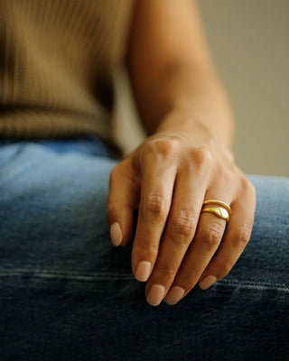 A close-up of a person's hand resting on their knee, wearing two slim gold bands rings - one signet and one band. The person is wearing an olive green ribbed knit sweater and blue denim jeans. A Rae Band stacked with an Oval Pinky Ring by George Rings, cast in solid 18K yellow gold.