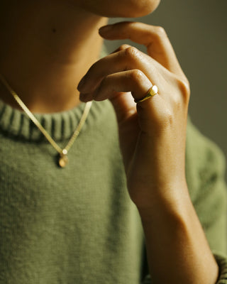 A close-up editorial photo of a woman wearing an Oval Pinky Ring by George Rings on her pinky finger, paired with a gold chain necklace and an olive green knit sweater, set against a dark, warm-toned background.