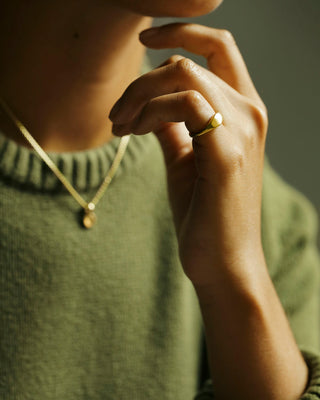 A close-up editorial photo of a woman wearing an Oval Pinky Ring by George Rings on her pinky finger, paired with a gold chain necklace and an olive green knit sweater, set against a dark, warm-toned background.