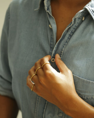 A close-up of a person wearing a light blue denim button-up shirt, with their hand adjusting the collar and button. The person is wearing several thin gold rings on their fingers. Rae Band by George Rings, cast in solid 18K yellow gold.