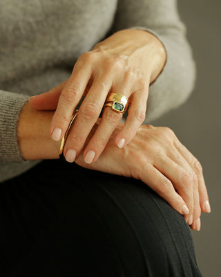 Close-up of hands with gold rings on a neutral background; blue-green tourmaline solitaire ring in solid 18K yellow gold by george rings 