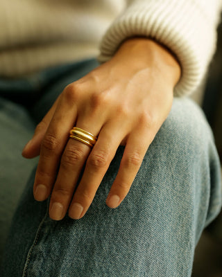 A close-up of a person's hand resting on their knee, wearing two slim gold bands rings. The person is wearing a cream ribbed knit sweater and blue denim jeans. Two Rae Bands by George Rings, cast in solid 18K yellow gold, stacked on either side of a Dominus Band Grand.