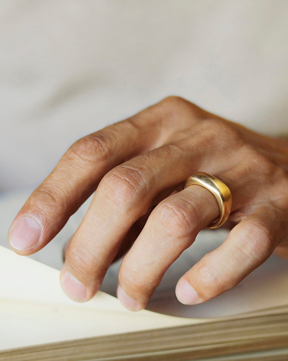 White man's hand turning the page of a book, wearing a solid 18k yellow gold heavy wedding ban on his ring finger. Noble Ring by George Rings for men, women, and all genders.