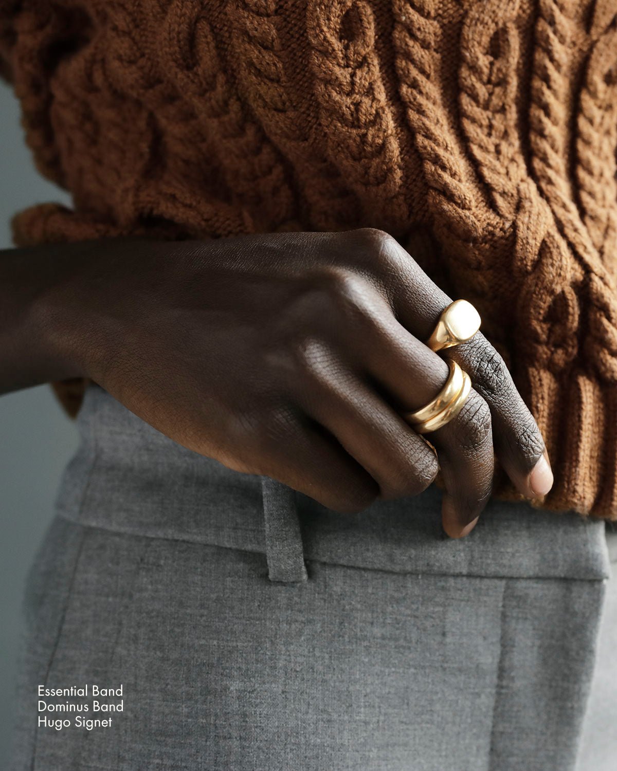 Woman holds touches her waist and shows three solid 18k yellow gold rings. She wears a solid 18k yellow gold heavy signet ring on her index finger. Hugo Signet Ring by George Rings. Essential Grande Band and Essential Band rings on her middle finger.