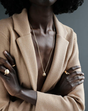 Black woman wears wool camel blazer with solid yellow gold rings and solid 18k yellow gold pillow pendant on a solid 14k yellow gold box chain. Pillow Pendant by George Rings.