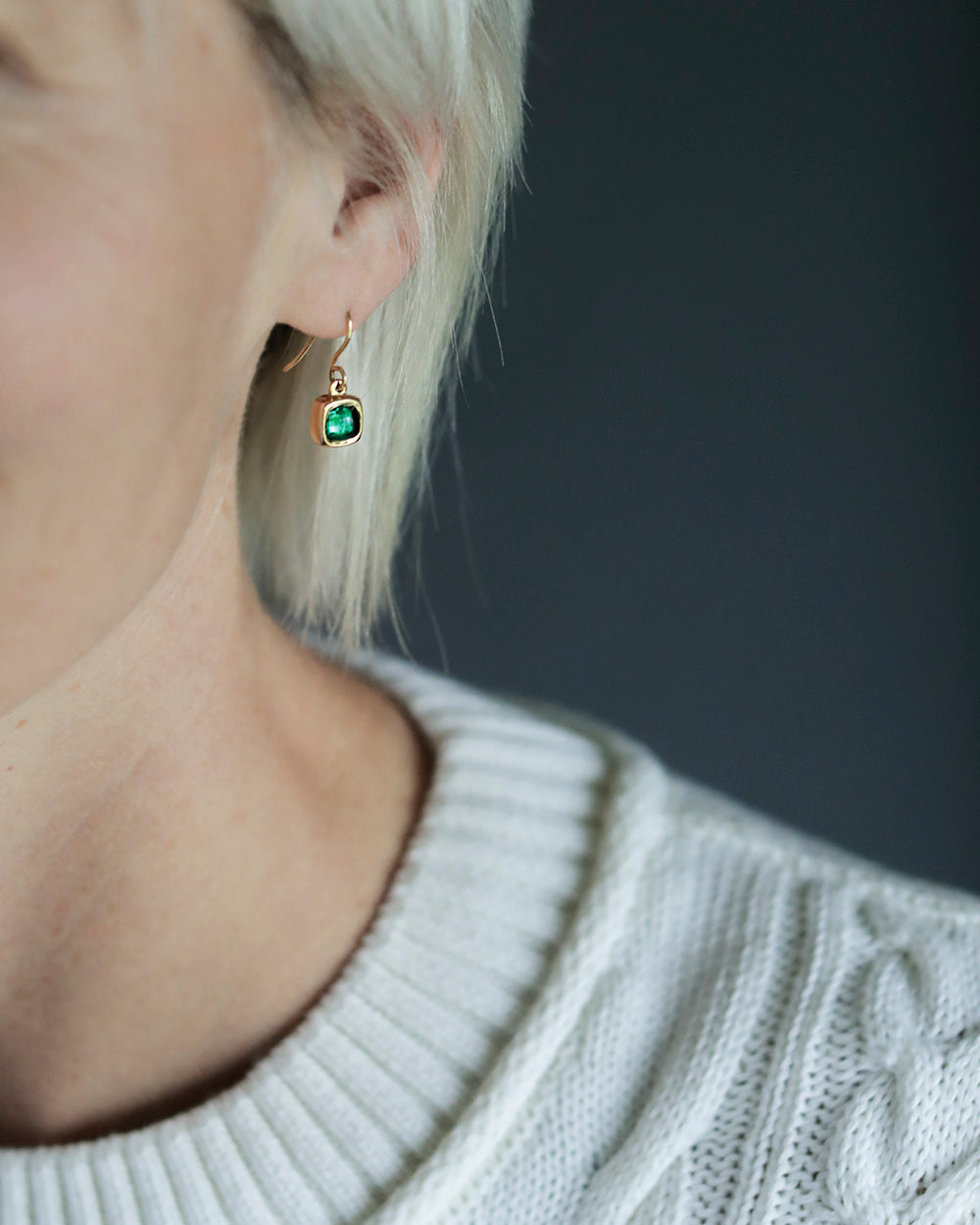 White woman wears a pair of green emerald rounded square gems inside solid 18k yellow gold bezels hanging from gold french wires.